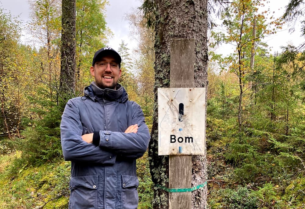 Niels Bom, wearing a warm blue winter coat and a blue baseball cap. He's standing in a Swedish forest with his arms crossed. He's standing next to a tree that has a sign on it. The sign is white and looks a bit worn. In black letters the sign say 'Bom'.